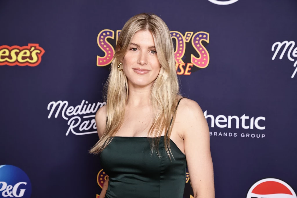 Eugenie Bouchard Młoda kobieta o jasnych, blond włosach ubrana w ciemnozieloną sukienkę stoi na tle ścianki z logotypami znanych marek i wydarzenia, uśmiecha się delikatnie, jej włosy opadają na ramiona.
