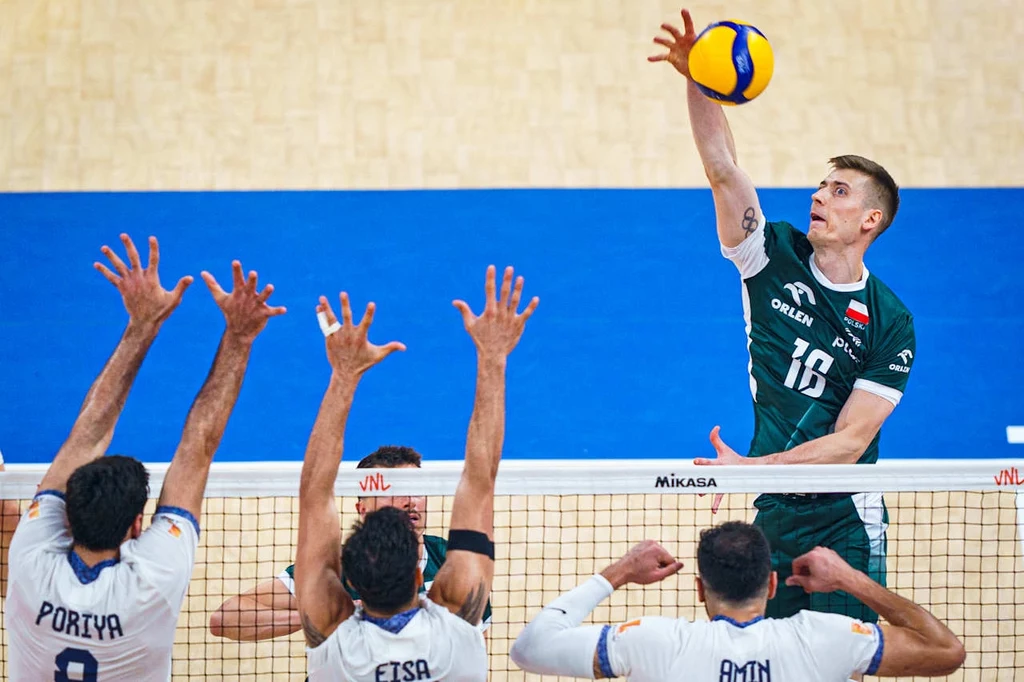 Liga Narodów siatkarzy. Kamil Semeniuk w trakcie meczu Polska - Iran Zawodnik w zielonym stroju wykonuje atak piłką siatkową ponad siatką, podczas gdy trzech przeciwników w białych koszulkach blokuje po drugiej stronie boiska. W tle jasnoniebieska powierzchnia boiska.