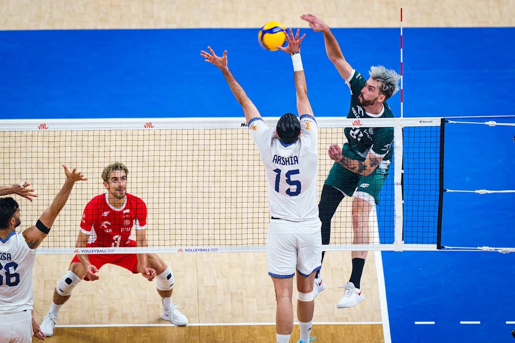 Tomasz Fornal w ataku Zawodnik w zielonym stroju atakuje piłkę podczas meczu siatkówki, podczas gdy gracz z numerem 15 w białym stroju próbuje zablokować atak przy siatce. Pozostali zawodnicy obserwują akcję z bliska. Tło stanowi niebiesko-beżowa podłoga hali sportowej.
