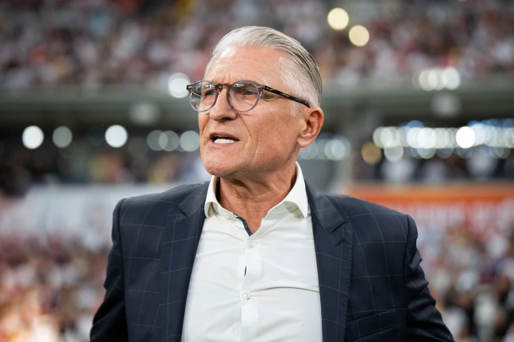 Adam Nawałka Mężczyzna w średnim wieku w eleganckim garniturze i białej koszuli, ma krótkie siwe włosy oraz okulary, stoi na tle rozmytego stadionu wypełnionego kibicami.