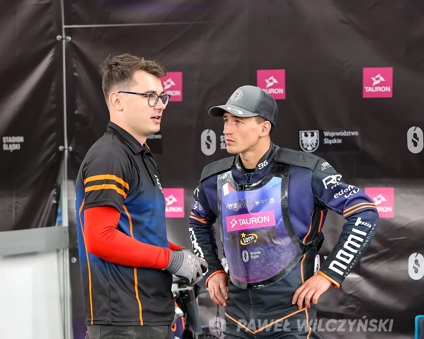 Dwóch mężczyzn rozmawia w strefie technicznej podczas sportowego wydarzenia. Jeden ubrany w sportowy strój, drugi w koszulkę i okulary, obaj wyglądają na skoncentrowanych. W tle widoczne logotypy sponsorów oraz Województwo Śląskie.