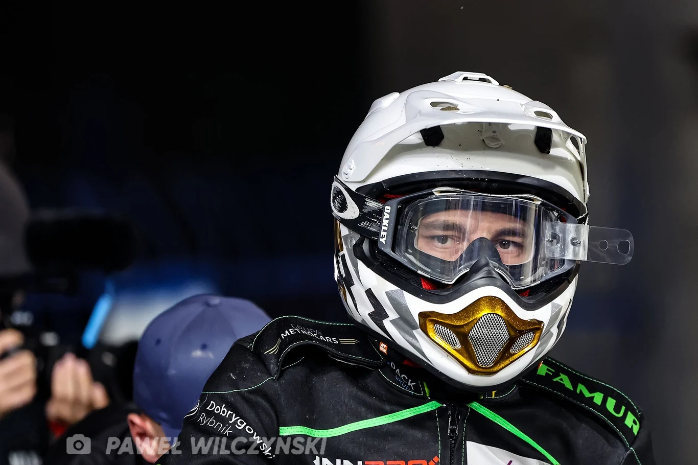 Jakub Jamróg Zawodnik ubrany w czarny kombinezon motocyklowy oraz biały kask z goglami, skoncentrowany przed rozpoczęciem wyścigu, w tle widoczne rozmyte postacie i sprzęt fotograficzny.