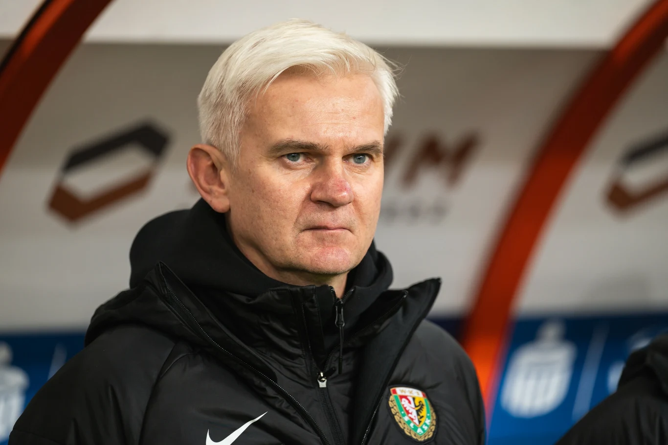 Mężczyzna o siwych włosach ubrany w czarną kurtkę sportową z widocznym herbem klubu piłkarskiego, stojący na tle ławki rezerwowych na stadionie.