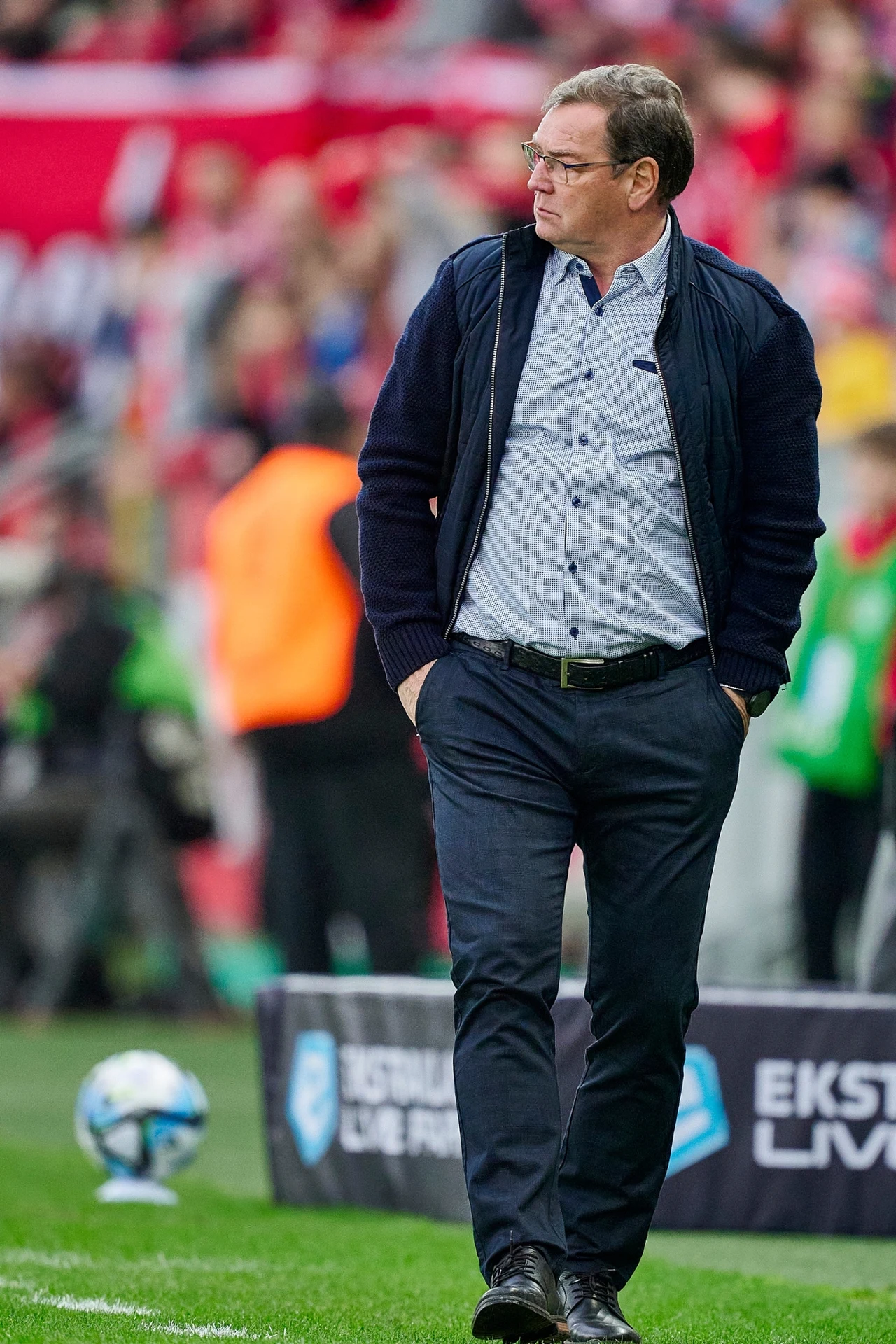 Mężczyzna w okularach ubrany w ciemną marynarkę i spodnie stoi przy linii bocznej boiska piłkarskiego, w tle rozmyci kibice i elementy stadionu oraz piłka.