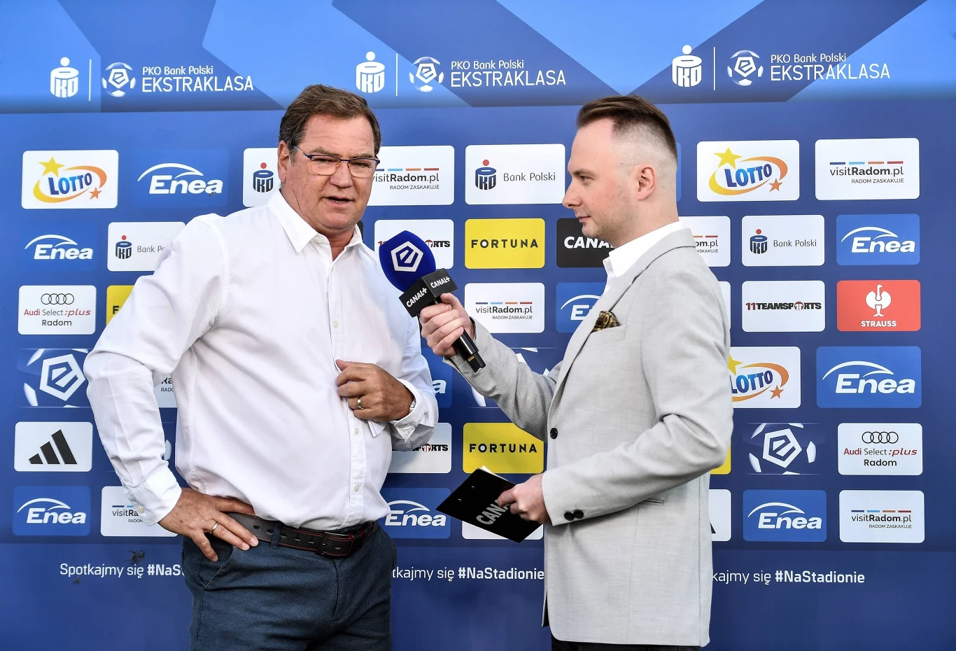 Dziennikarz w jasnej marynarce przeprowadza wywiad z mężczyzną w białej koszuli na tle ścianki z logo sponsorów i Ekstraklasy.