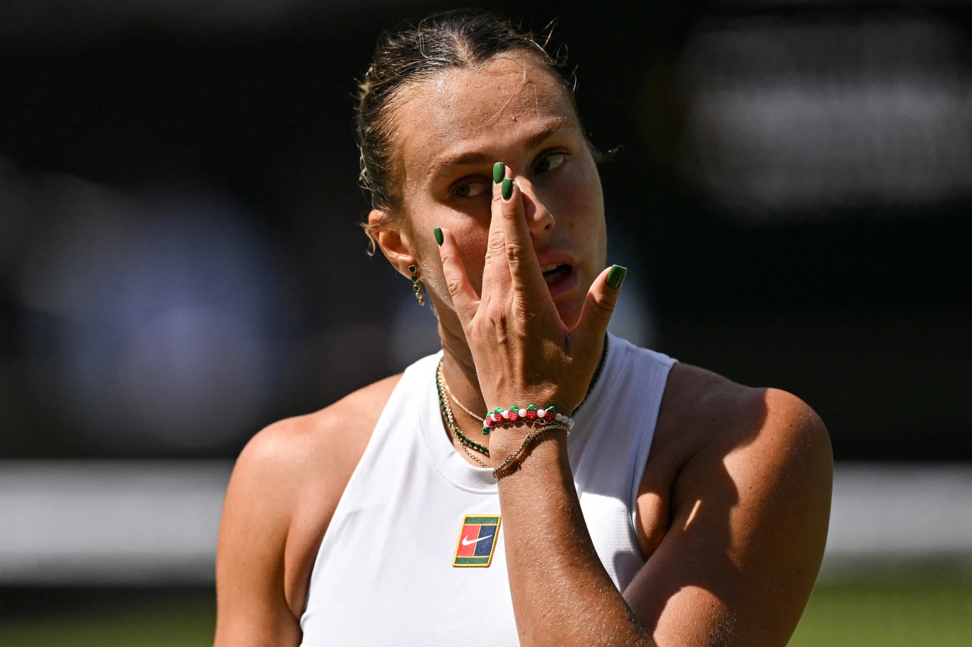 Aryna Sabalenka przegrała z Amandą Anisimovą w półfinale Wimbledonu Kobieta ubrana w biały sportowy strój przeciera oko dłonią, ma spięte włosy i kolorowy lakier na paznokciach, na szyi nosi bransoletki i naszyjnik, w tle rozmyte zielone otoczenie, prawdopodobnie kort tenisowy.