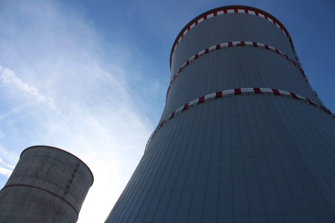 Dwie wieże chłodnicze elektrowni jądrowej w Ostrowcu. Obiekt leży kilka kilometrów od granicy z Litwą Dwie wielkie chłodnie kominowe elektrowni z charakterystycznymi czerwonymi pasami na górze, jedna mniejsza w tle po lewej stronie, błękitne niebo w tle, światło padające zza konstrukcji.