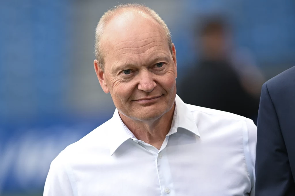 Mężczyzna w średnim wieku ubrany w białą koszulę z lekko uśmiechniętym wyrazem twarzy, na rozmytym tle z niebieskimi akcentami.