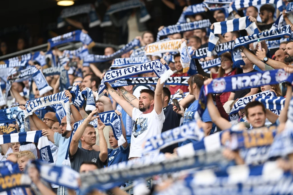 Tłum kibiców dopingujących swój zespół piłkarski Lech Poznań, uniesione w górę szaliki w klubowych barwach biało-niebieskich, atmosfera sportowego wsparcia i entuzjazmu.