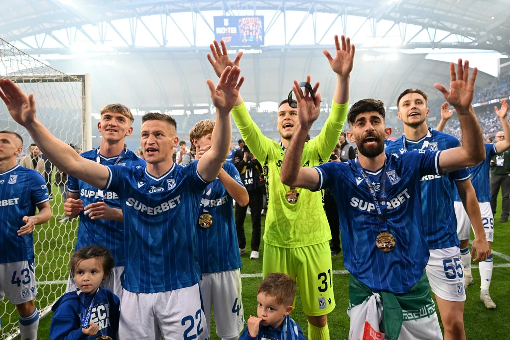 Piłkarze ubrani w niebieskie stroje świętują na murawie stadionu, unosząc ręce w górę i uśmiechając się do kibiców; w tle widać tłum ludzi na trybunach, wokół piłkarzy stoją dzieci.