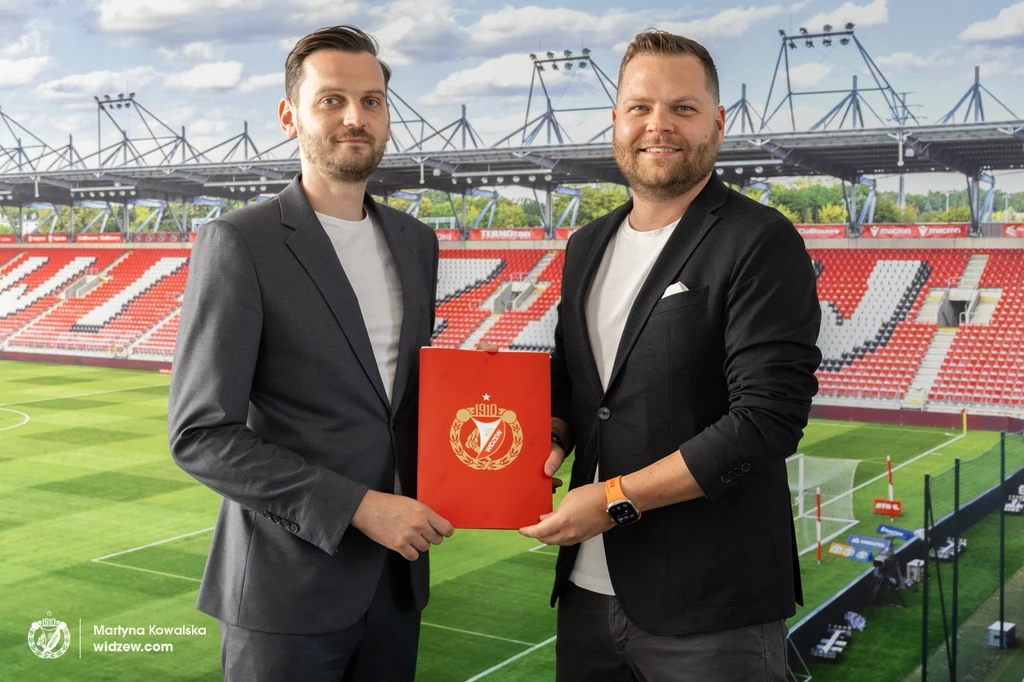 Piotr Urban i wiceprezes Widzewa Maciej Szymański Dwie elegancko ubrane osoby stojące na tle stadionu piłkarskiego, jedna z nich trzyma czerwony segregator z herbem klubu sportowego, widoczne trybuny i murawa stadionu w tle.