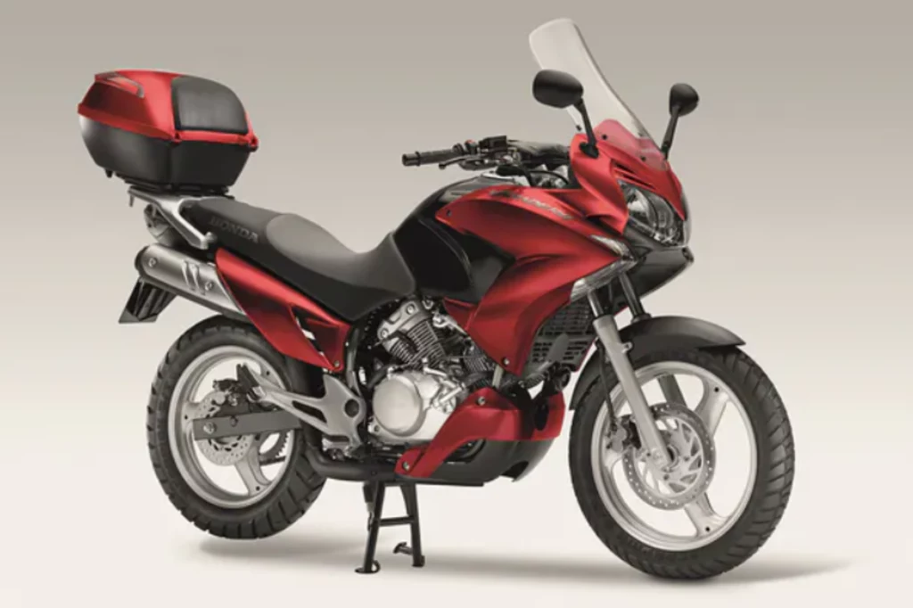 Czerwony motocykl turystyczny z kufrem i szybą, ustawiony na centralnej podnóżce, dobrze widoczne elementy takie jak hamulec tarczowy i układ wydechowy.