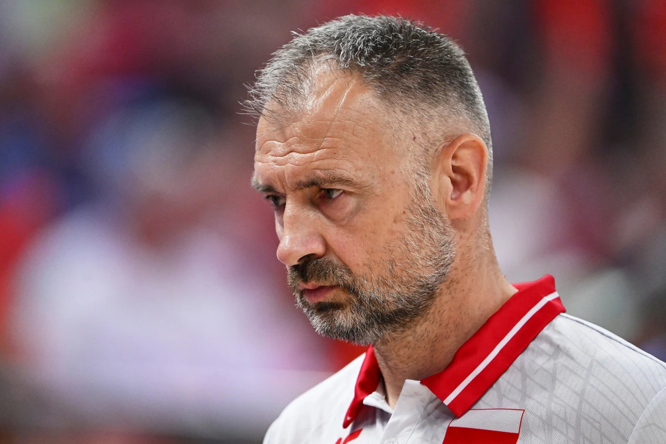Dojrzały mężczyzna o krótkich, siwiejących włosach i brodzie, ubrany w sportową koszulkę z czerwonym kołnierzykiem i białymi oraz czerwonymi elementami, na twarzy poważny wyraz skupienia. W tle niewyraźne sylwetki i czerwone odcienie, sugerujące obecno...