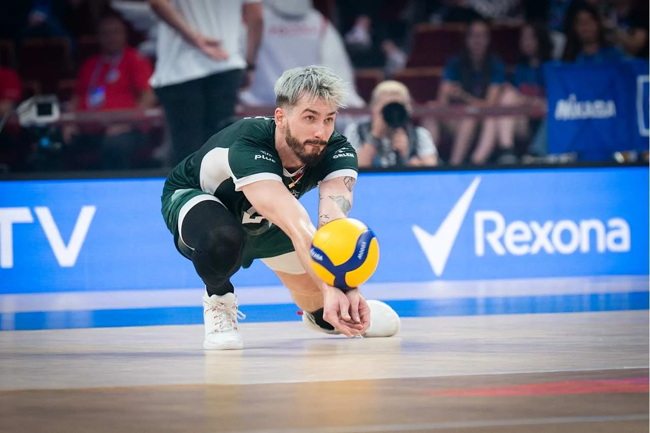 Zawodnik siatkówki w momencie odbioru piłki podczas intensywnego meczu, dynamiczna poza, sportowe emocje na hali, widoczne logo sponsora na bandzie oraz skupienie na rozgrywce.