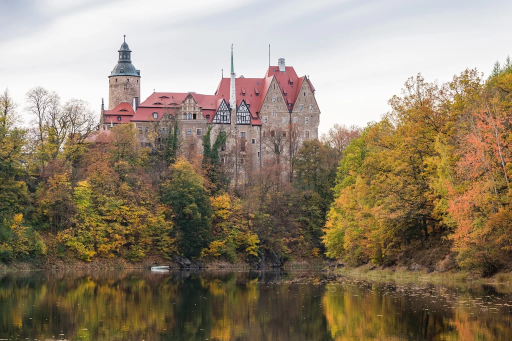 Zamek z czerwonymi dachami położony na wzgórzu wśród kolorowych, jesiennych drzew nad spokojną powierzchnią jeziora odbijającą budowlę i otoczenie.