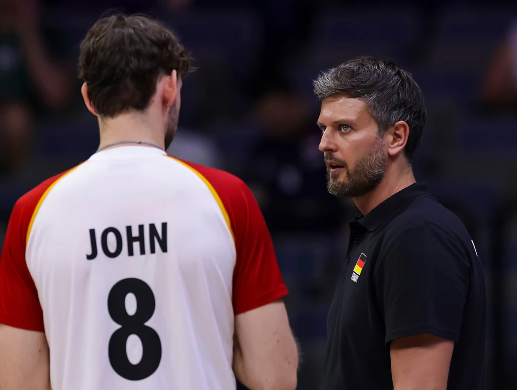 Michał Winiarski i Filip John Siatkarz w koszulce z nazwiskiem John i numerem 8 stoi twarzą do trenera ubranym na czarno, prowadząc rozmowę podczas meczu siatkówki.