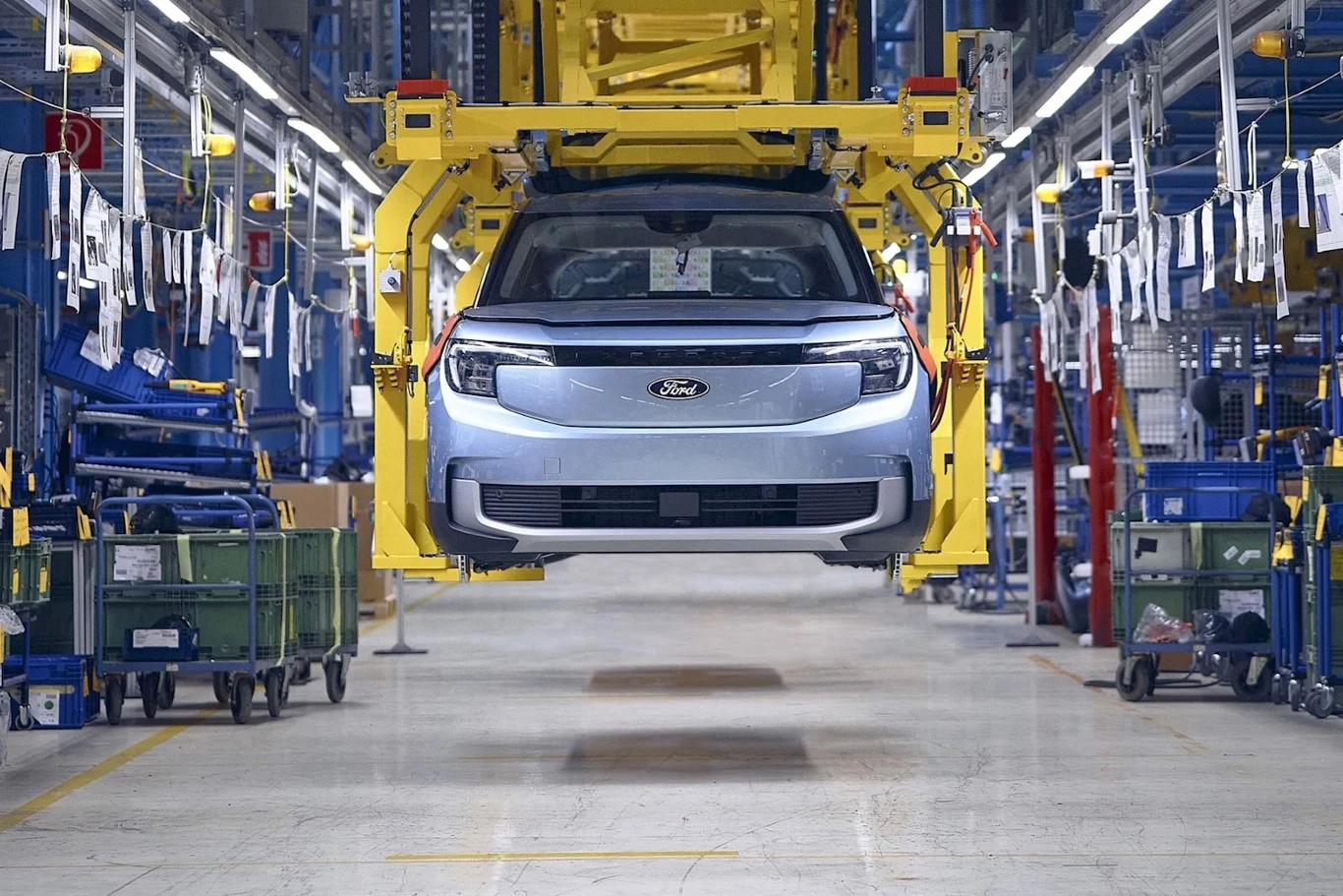 Ford zgodził się na program dobrowolnych odejść z fabryki w Kolonii. Do 2032 roku z zakładu odejdzie 2900 pracowników Auto osobowe marki Ford zawieszone na żółtej ramie w trakcie montażu na linii produkcyjnej w nowoczesnej fabryce oświetlonej jasnym światłem, wokół widoczne pojemniki z częściami i zawieszone taśmy produkcyjne.