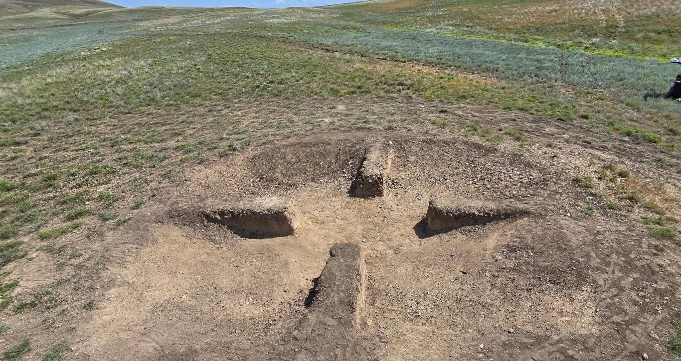 Duży wykop ziemny w kształcie krzyża z widocznymi wyraźnymi krawędziami na otwartym, pagórkowatym terenie porośniętym trawą; w oddali rozciąga się pusta przestrzeń bez zabudowań.