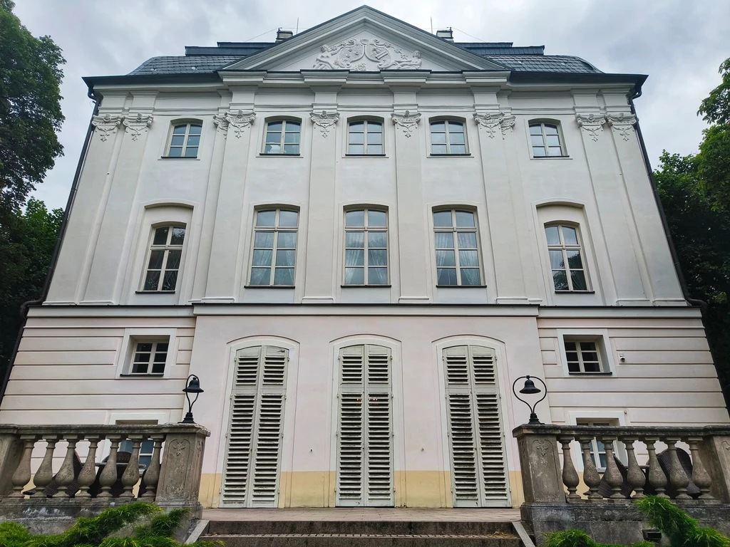 Zamierzasz odwiedzić województwo kujawsko-pomorskie? Ostromecko to obowiązkowy przystanek Fasada eleganckiej, klasycystycznej kamienicy z białymi ścianami, dużymi oknami i balkonem otoczonym balustradą, po bokach lampy ogrodowe, budynek otaczają drzewa.