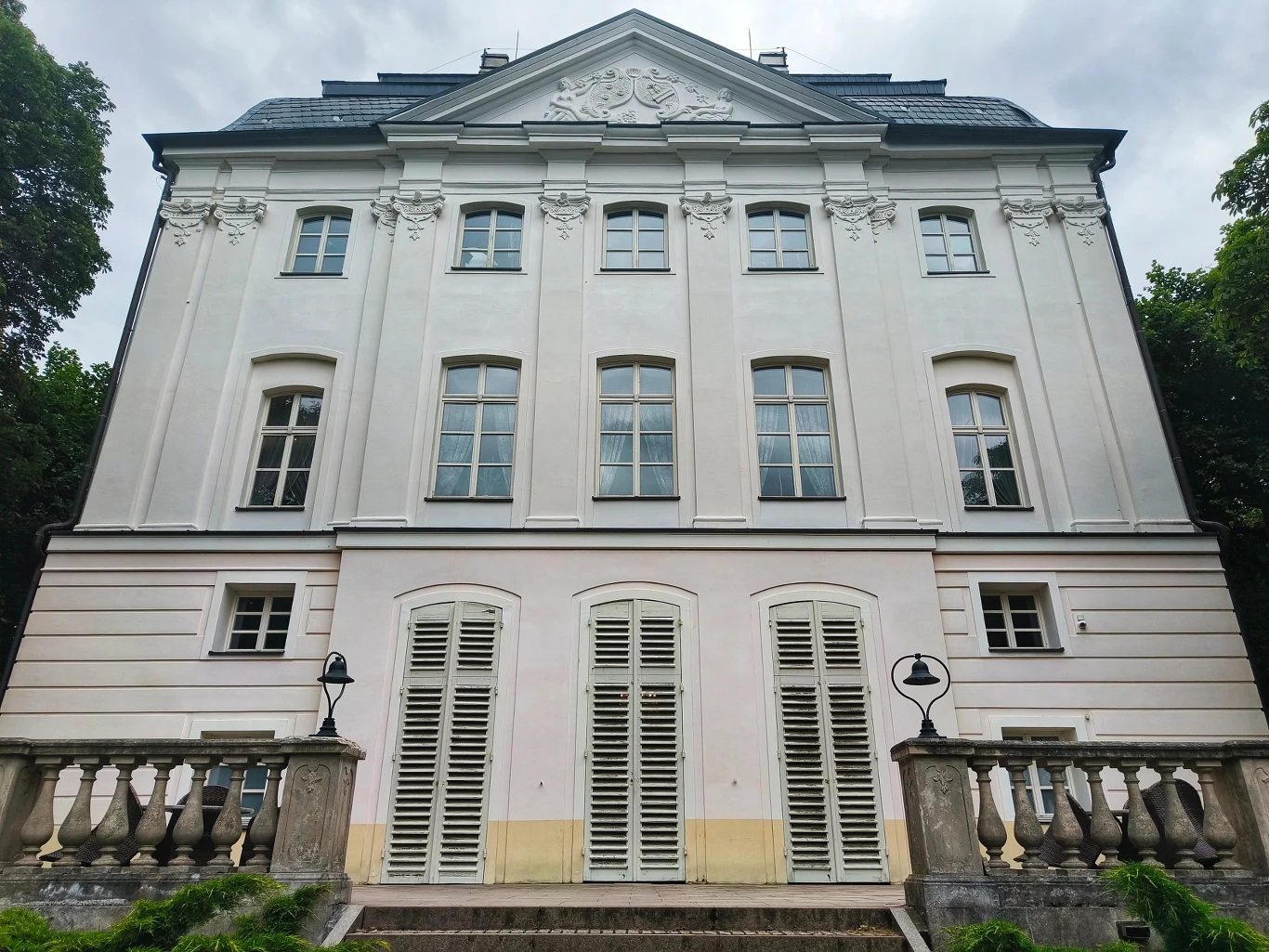 Zamierzasz odwiedzić województwo kujawsko-pomorskie? Ostromecko to obowiązkowy przystanek Fasada eleganckiej, klasycystycznej kamienicy z białymi ścianami, dużymi oknami i balkonem otoczonym balustradą, po bokach lampy ogrodowe, budynek otaczają drzewa.