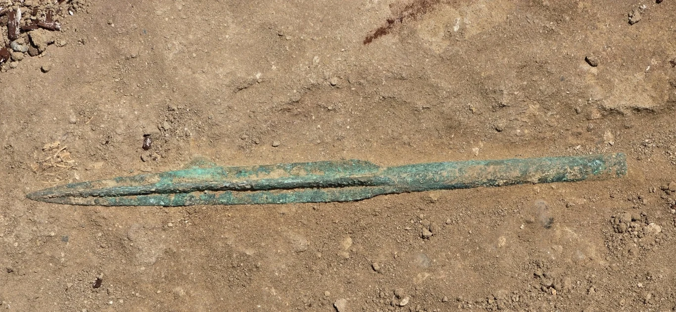 Starożytne ostrze wykonane z metalu częściowo pokryte patyną, leżące na ziemistym podłożu wykopaliskowym.