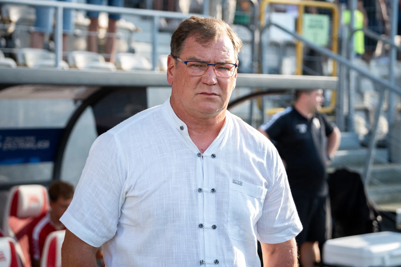 Jan Urban Mężczyzna w okularach ubrany w jasną koszulę stoi na stadionie, w tle widoczne są ławki rezerwowych oraz inni ludzie.