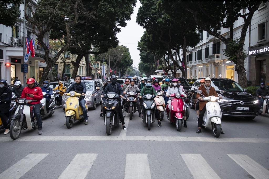 Grupa osób na skuterach i motocyklach czeka na zielone światło przed przejściem dla pieszych, po obu stronach ulicy widoczne są drzewa oraz zabudowania miejskie.