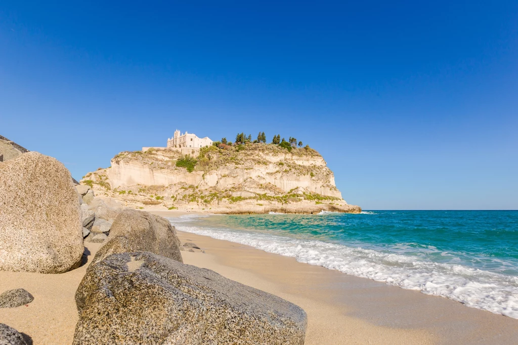 Tropea w Kalabrii to bez wątpienia jedno z najpiękniejszych miast na południu Włoch Piaszczysta plaża z dużymi głazami, niebieskie morze oraz malownicza skała z budowlą na szczycie i jasnoniebieskim, bezchmurnym niebem w tle.