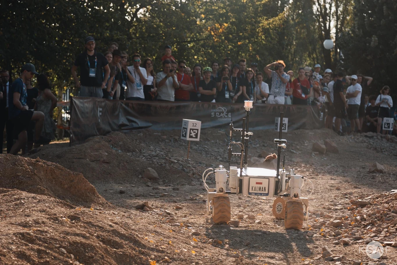 European Rover Challenge 2025 na AGH. Kraków będzie stolicą robotyki. Robot terenowy poruszający się po piaszczystym, kamienistym torze konkursowym, otoczony przez obserwującą publiczność. W tle zgrupowanie ludzi, numeracja stanowisk i drzewa podkreślające plenerowy charakter wydarzenia.