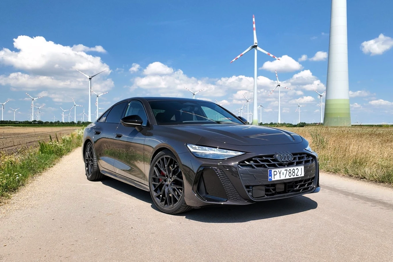 Nowe Audi A6 z hybrydą plug-in pokazuje, że nie tylko diesle są dziś oszczędne. Czarny samochód marki Audi stoi na polnej drodze, w tle liczne wiatraki oraz błękitne niebo z pojedynczymi chmurami.