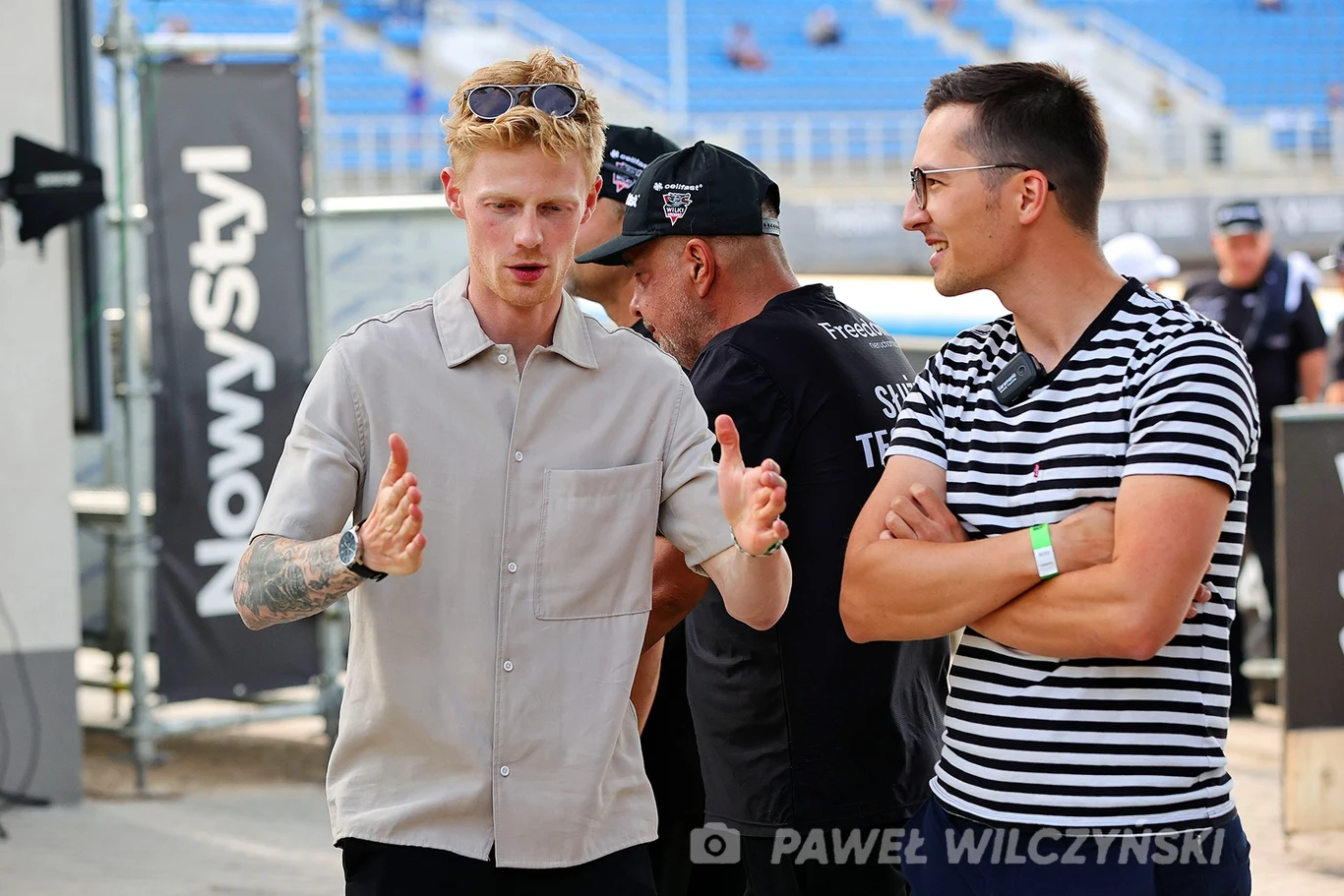 Dwóch młodych mężczyzn rozmawia ze sobą na tle niebieskich trybun stadionu, jeden gestykuluje, drugi ma skrzyżowane ramiona; w tle widocznych jest kilku innych ludzi, część z nich zwrócona tyłem.