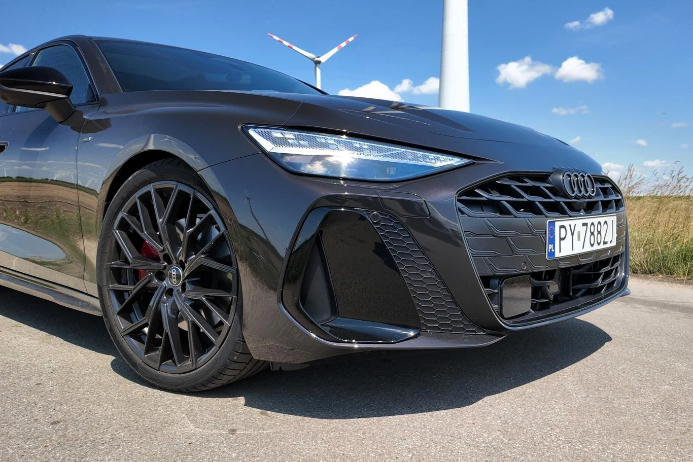 Spalinowe Audi A6 różni się od elektrycznego m.in. niezabudowaną osłoną chłodnicy, ale też innymi światłami. Luksusowy czarny samochód marki Audi zaparkowany na asfalcie, z widocznym dużym grillem, nowoczesnym designem reflektorów oraz sportowymi felgami; w tle przydrożne pole i fragment turbiny wiatrowej na tle nieba z chmurami.