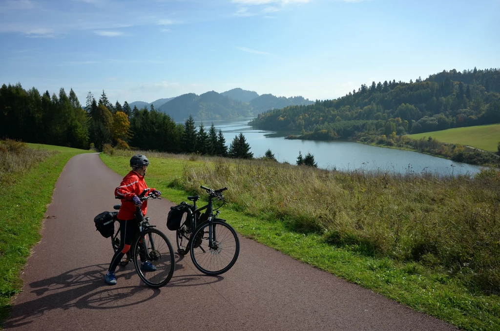 Osoba w kasku rowerowym na drodze rowerowej z dwoma rowerami, otoczona zielonymi wzgórzami i lasem, z widokiem na rzekę lub jezioro oraz górski krajobraz w tle.