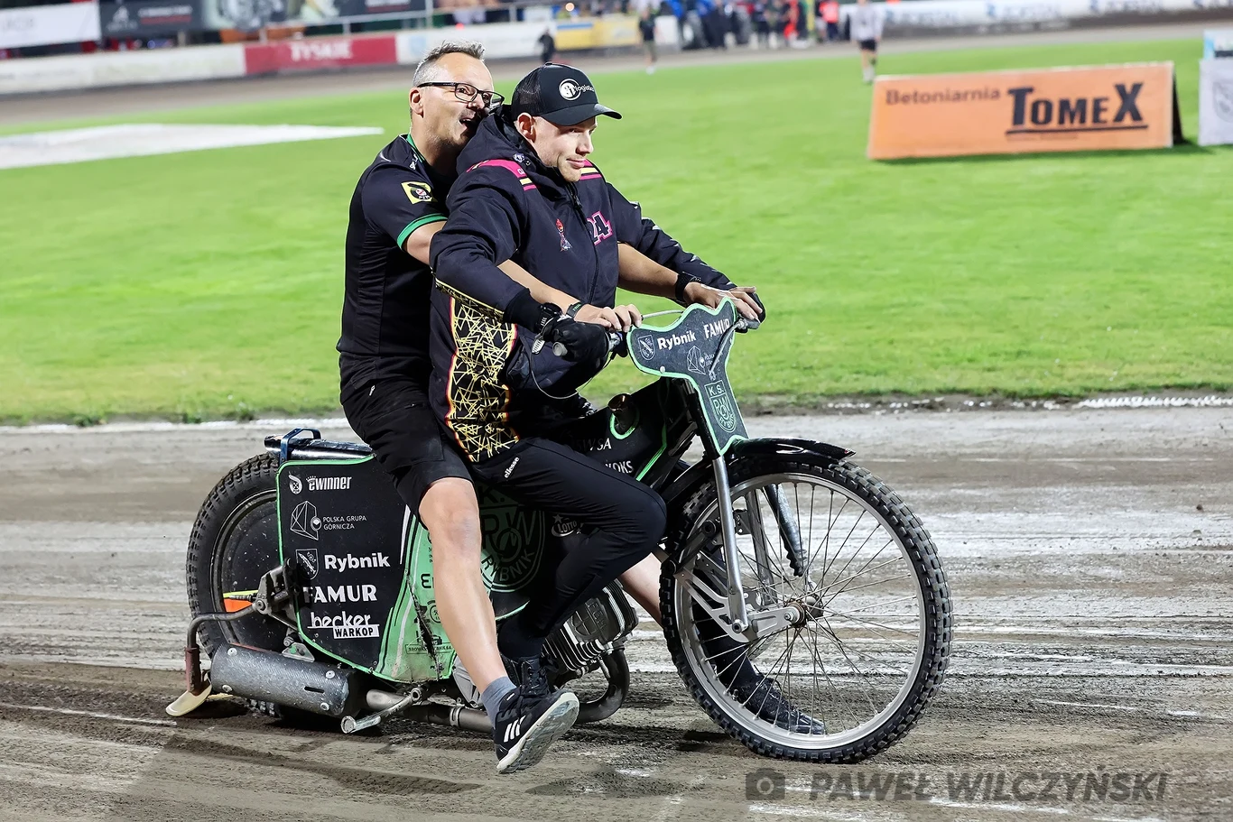 Patrick Hansen na motocyklu, tuż po okropnej kontuzji Dwóch mężczyzn jedzie na żużlowym motocyklu po torze, jeden z nich ubrany jest w strój zawodnika, drugi w sportową odzież, na trybunach widoczni są kibice, w tle banery reklamowe.