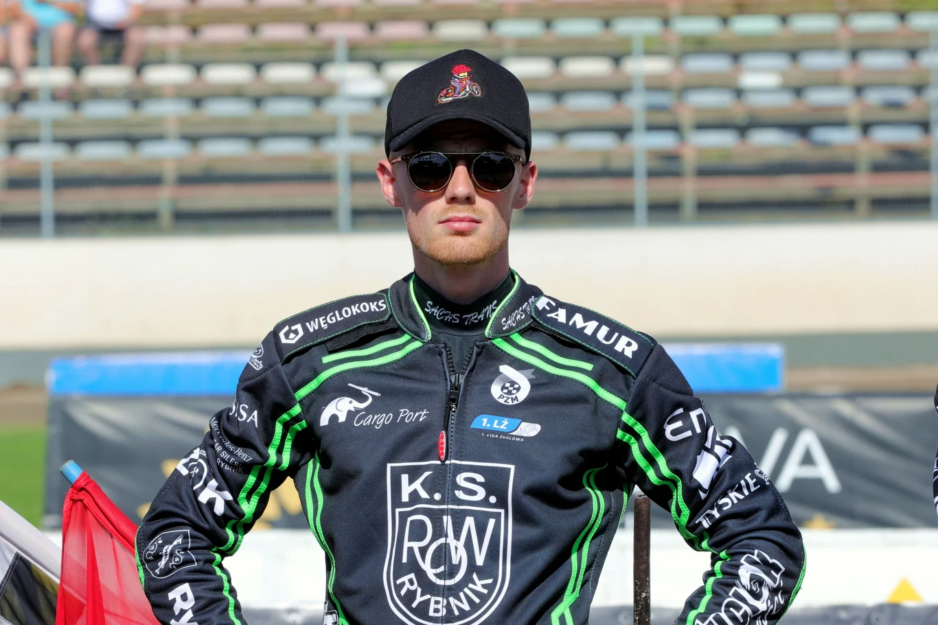 Patrick Hansen w sezonie 2023, w barwach ROW-u Rybnik Mężczyzna w ciemnych okularach i czapce z daszkiem stoi w stroju żużlowym z logo K.S. ROW Rybnik na tle trybun stadionu.