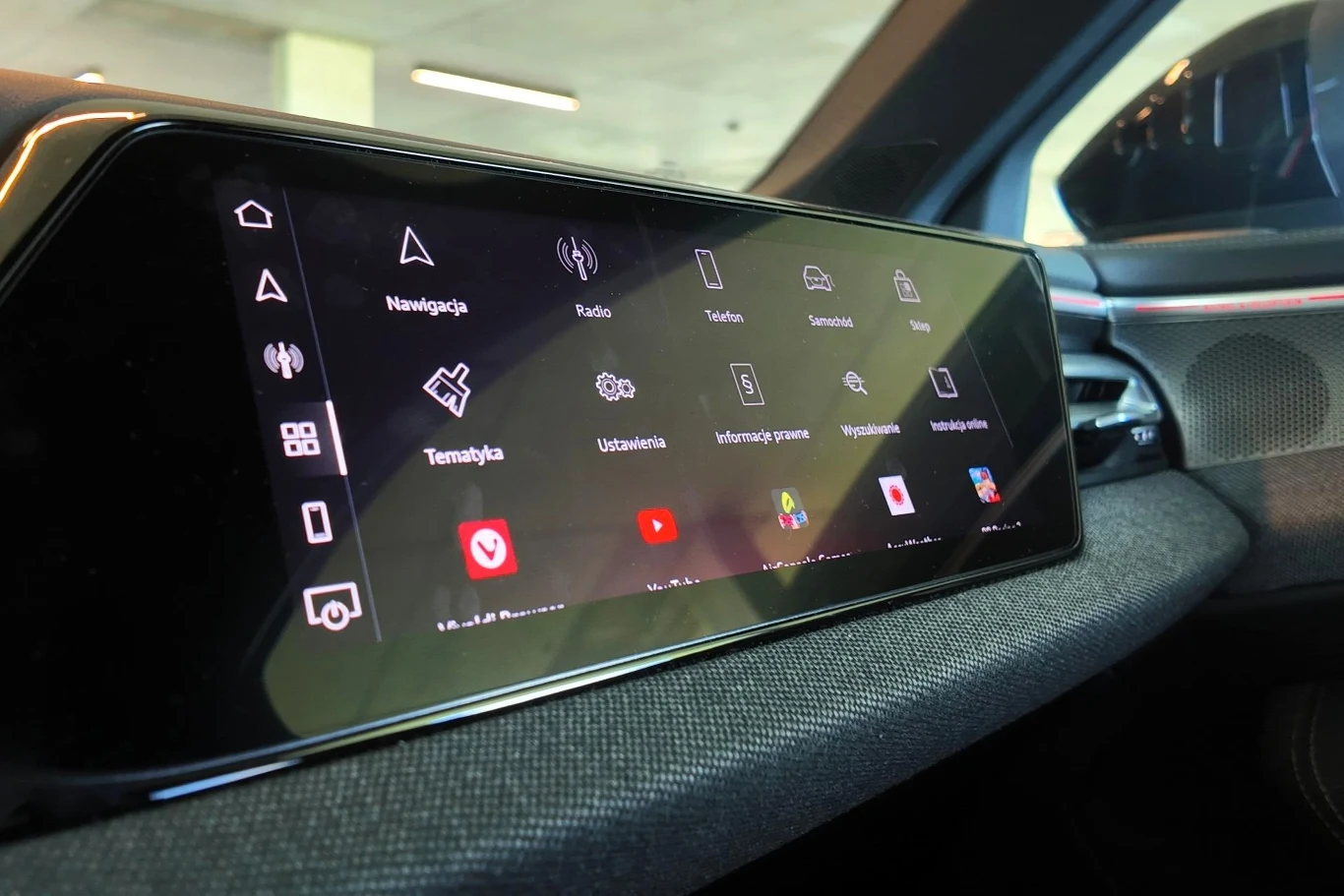 Audi A6 opcjonalnie może być wyposażone w ekran znajdujący się przed kierowcą. Duży ekran dotykowy systemu multimedialnego samochodu z widocznymi ikonami nawigacji, radia, ustawień oraz aplikacji, w nowoczesnym wnętrzu auta.