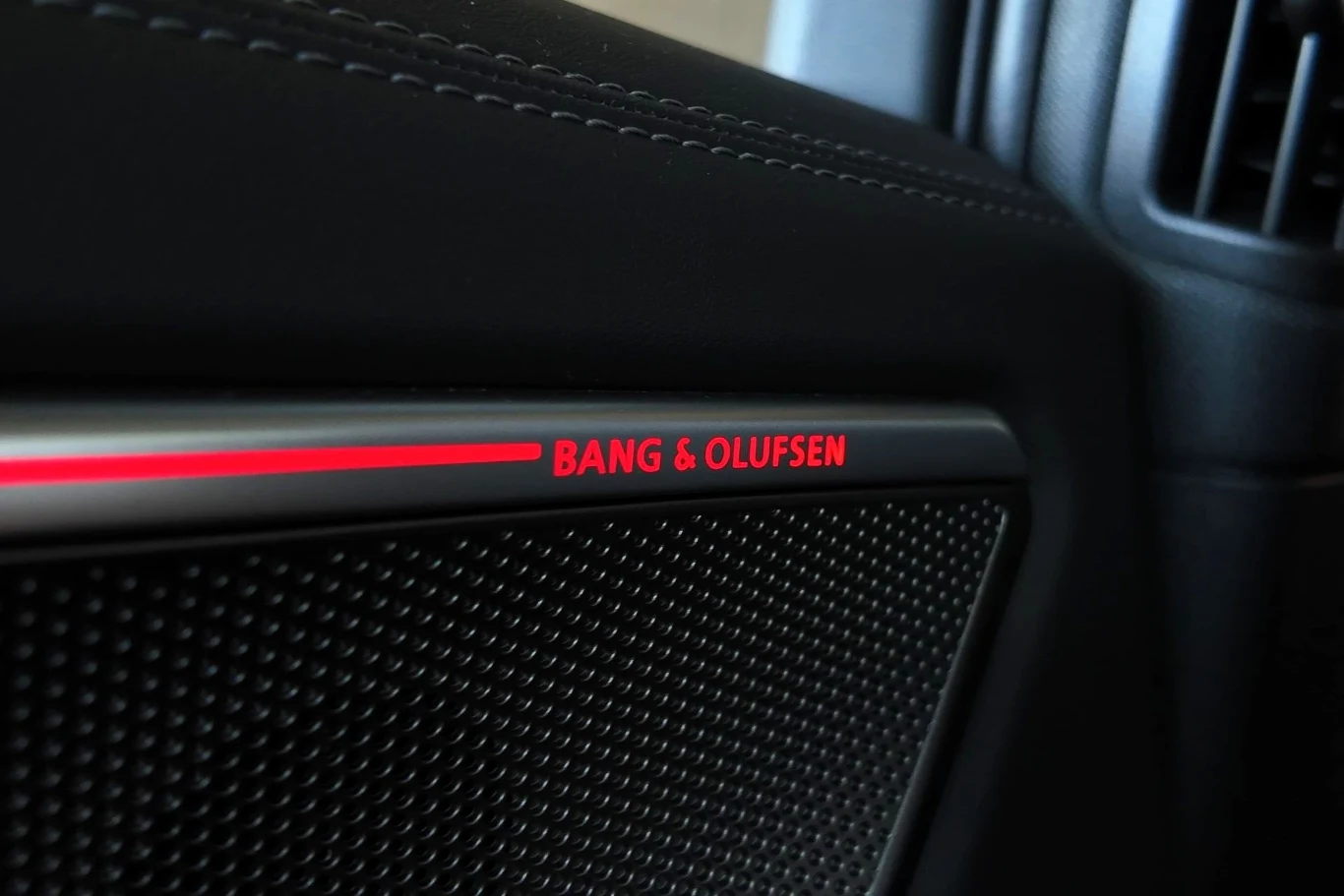 Opcjonalnie Audi A6 może być wyposażone w 16- lub 20-głośnikowe nagłośnienie od firmy Bang & Olufsen. Czarna kratka głośnika z napisem Bang & Olufsen podświetlonym na czerwono, element wnętrza samochodu w luksusowym stylu.