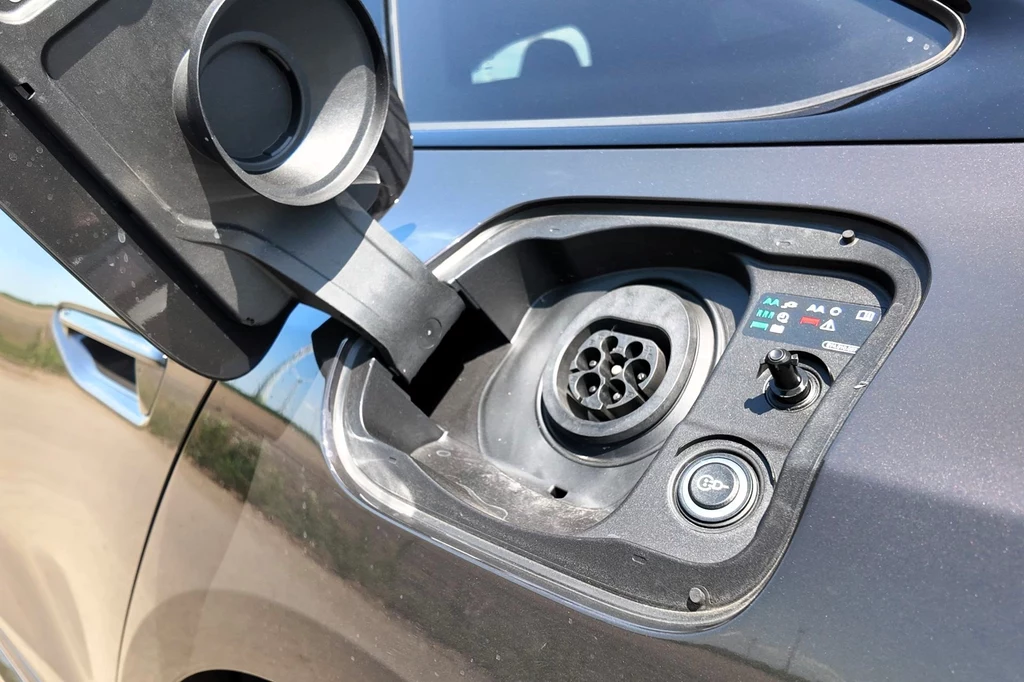 Gniazdo ładowania samochodu elektrycznego z otwartą klapką, widoczne szczegóły złącza oraz przycisk otwierania pokrywy, częściowo widoczna karoseria pojazdu.