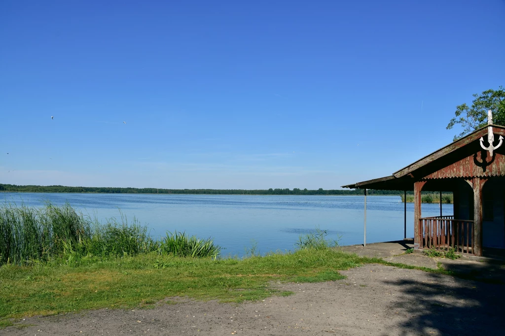 Domek letniskowy z drewnianą werandą nad brzegiem spokojnego jeziora, wokół gęsta roślinność i trawa, błękitne niebo bez chmur, cisza i spokój natury.
