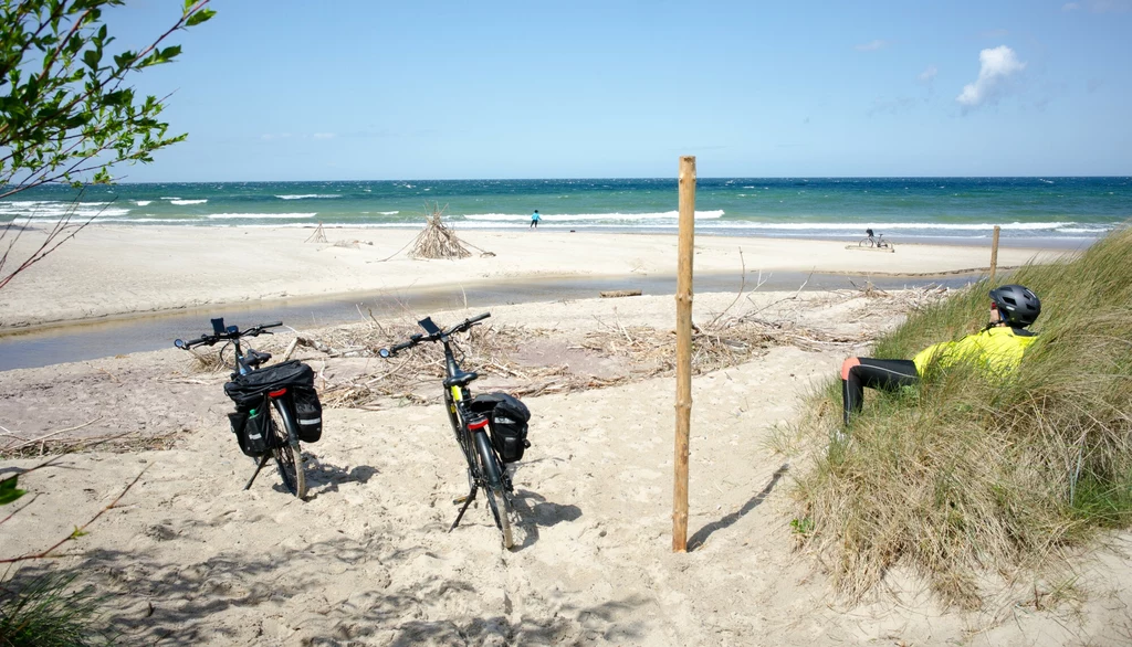 Dwa rowery stojące na piaszczystej plaży, obok siedzi osoba w żółtej kurtce i kasku rowerowym, w tle falujące morze i błękitne niebo