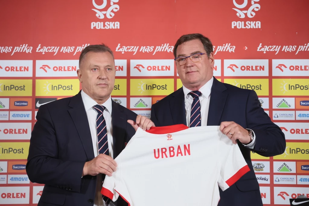 Cezary Kulesza, Jan Urban Dwóch mężczyzn w garniturach trzyma koszulkę piłkarską z napisem 'URBAN', stojąc przed ścianą sponsorów z logotypami firm oraz herbem Polski.