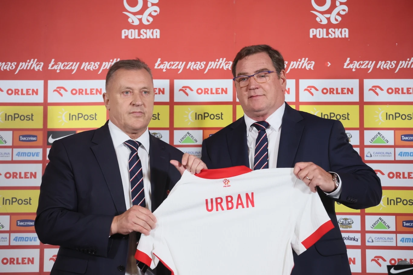 Dwóch mężczyzn w garniturach trzyma koszulkę piłkarską z napisem 'URBAN', stojąc przed ścianą sponsorów z logotypami firm oraz herbem Polski.