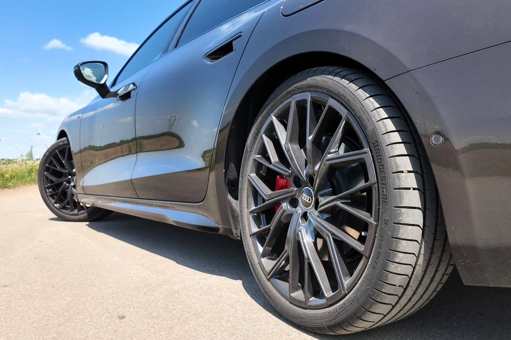 Czarny sportowy samochód marki Audi zaparkowany na asfalcie, z wyeksponowanymi aluminiowymi felgami i niskoprofilowymi oponami, w tle jasnoniebieskie niebo z chmurami.
