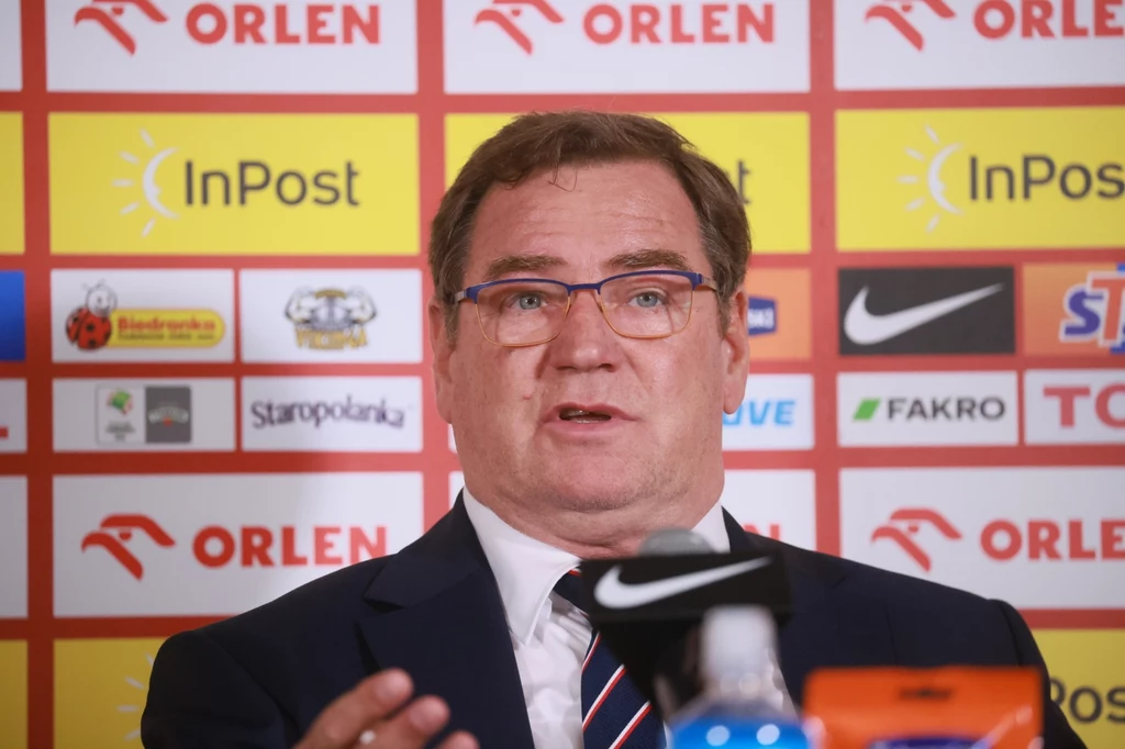 Mężczyzna w garniturze i okularach siedzi na tle ściany z logotypami sponsorów podczas konferencji prasowej, wyciąga dłoń w geście podkreślającym wypowiedź.