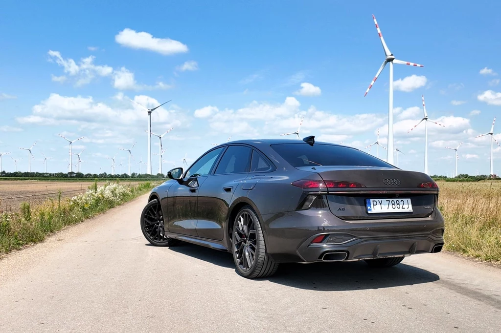 Czarny samochód osobowy marki Audi zaparkowany na wiejskiej drodze asfaltowej, w tle widoczne liczne turbiny wiatrowe oraz niebieskie niebo z kilkoma chmurami.