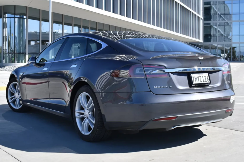 Szary samochód elektryczny marki Tesla Model S zaparkowany na tle nowoczesnego budynku ze szklaną fasadą, w słoneczny dzień, widoczna tylna część pojazdu z kalifornijską tablicą rejestracyjną.