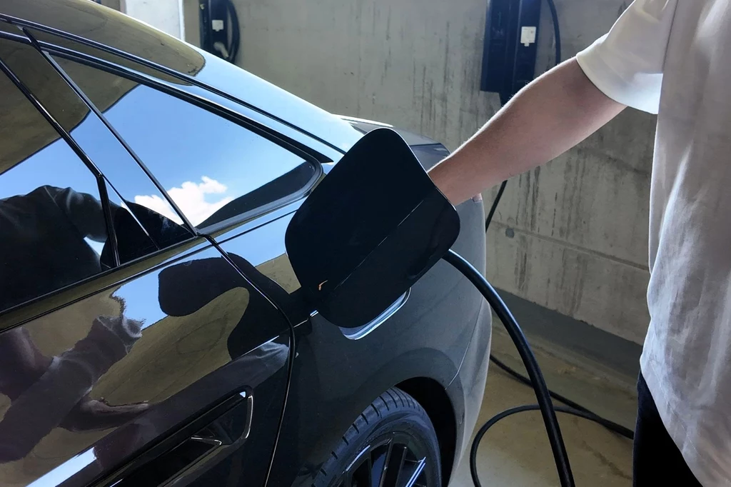 Nowoczesny samochód elektryczny podczas ładowania przy stacji ładowania, widoczna ręka osoby podłączającej kabel do portu ładowania pojazdu.