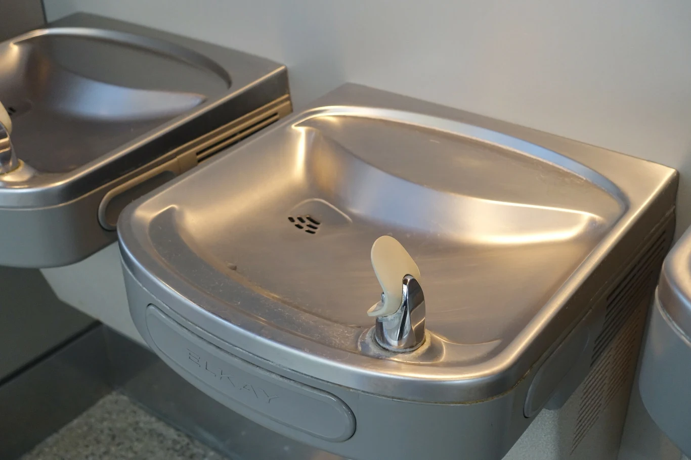 Srebrna fontanna wodna wbudowana w ścianę, z przyciskiem do uruchamiania strumienia wody, obok znajdują się inne podobne urządzenia.