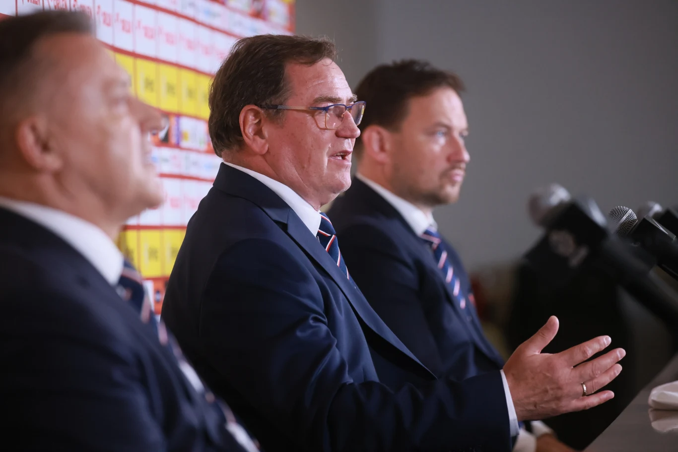 Trzech mężczyzn w garniturach siedzi przy stole konferencyjnym, jeden z nich przemawia do mikrofonu, w tle widoczna tablica z logotypami sponsorskimi.