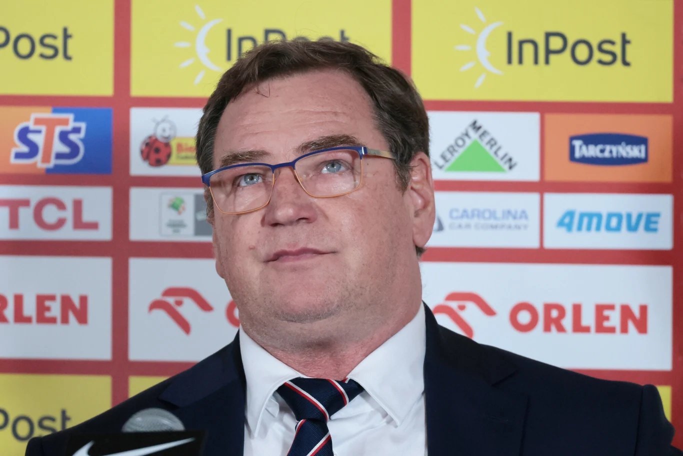 Mężczyzna w garniturze i okularach stoi na tle ściany z logotypami sponsorów, wygląda poważnie, jest w trakcie konferencji prasowej lub oficjalnego wystąpienia.